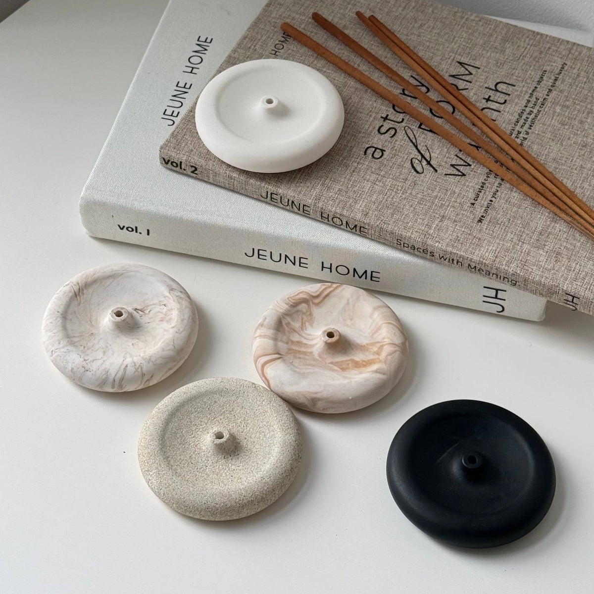 Four round stone incense burners in white, beige, and black, displayed on a white surface next to a book and wooden incense sticks.
