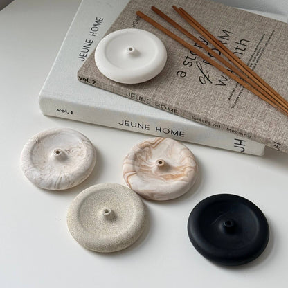 Four round stone incense burners in white, beige, and black, displayed on a white surface next to a book and wooden incense sticks.
