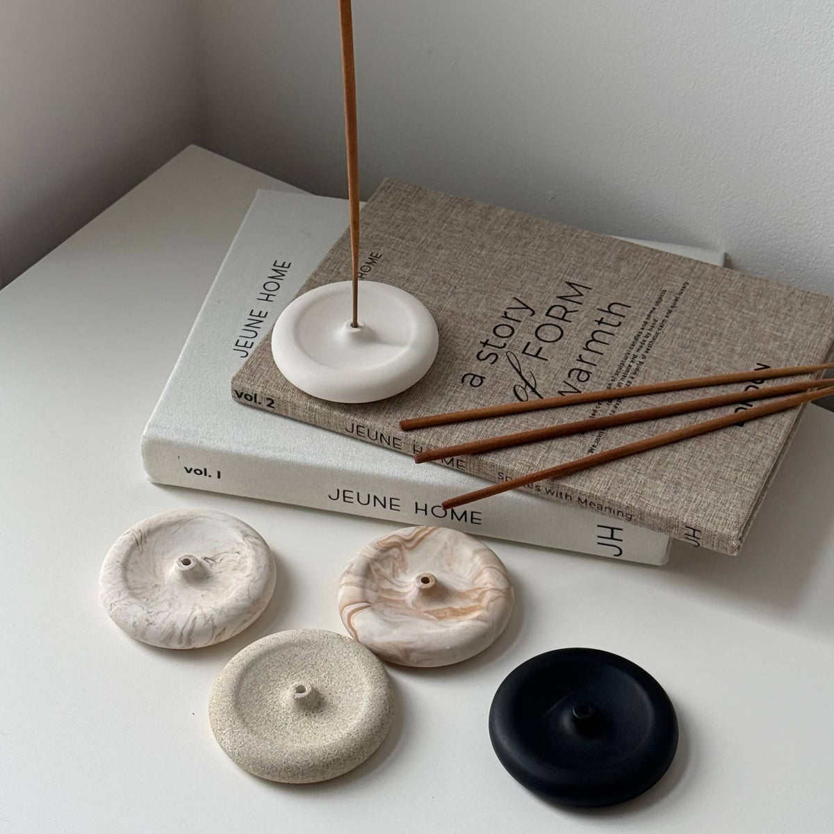 Four round incense burners in white, beige, and black are arranged on a white surface next to a book and incense sticks. The scene is styled in a minimalist, clean interior setting.