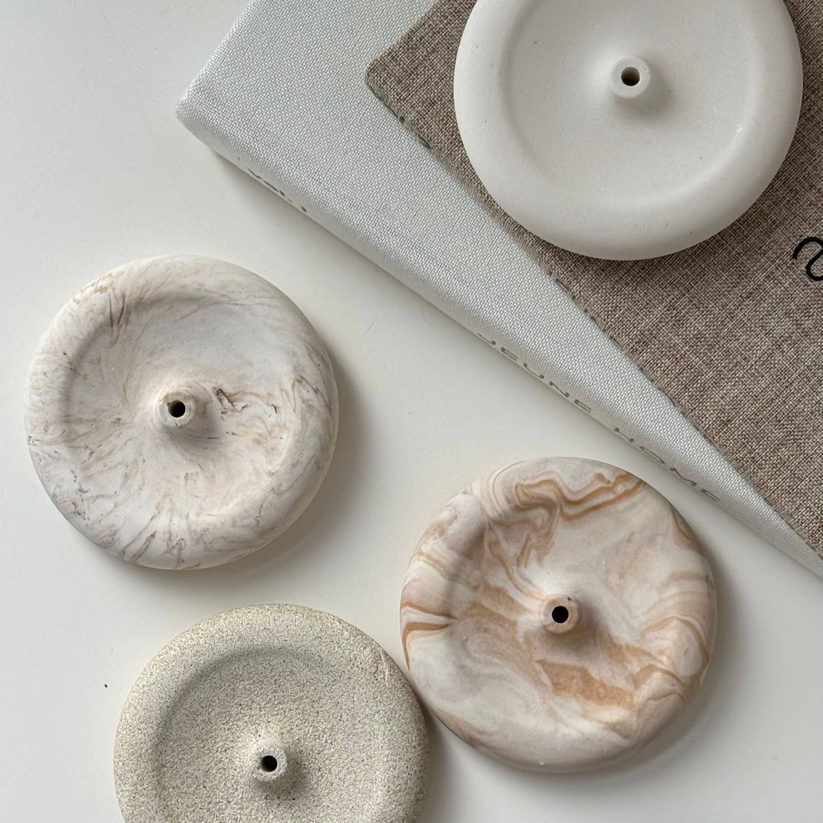 Four round ceramic candle holders with different textures and colors, including white, beige, marbled cream, and matte black, arranged on a light surface next to a book.
