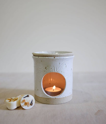 A ceramic candle warmer with a lid and a lit tea light candle inside, shown on a light wooden surface with two small wax melts in the foreground against a neutral background.