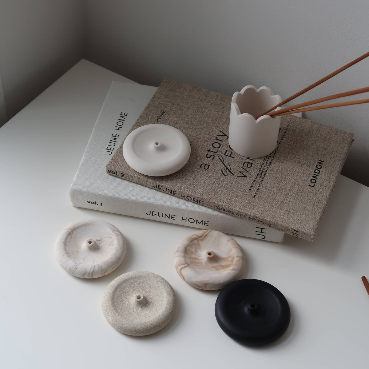 Four round ceramic candle holders in cream, beige marble, and matte black, arranged on a white tabletop next to a linen-covered book and a small white vessel holding wooden sticks.