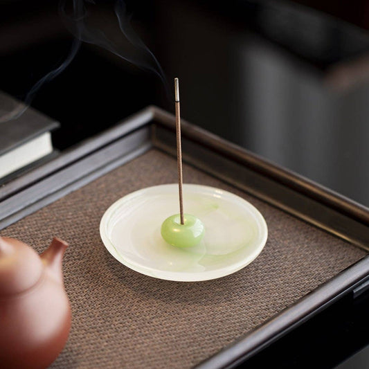 A close-up of a Gohobi London incense holder featuring a small white dish with a green incense ball and a smoking incense stick, styled on a dark tray next to a terracotta teapot.
