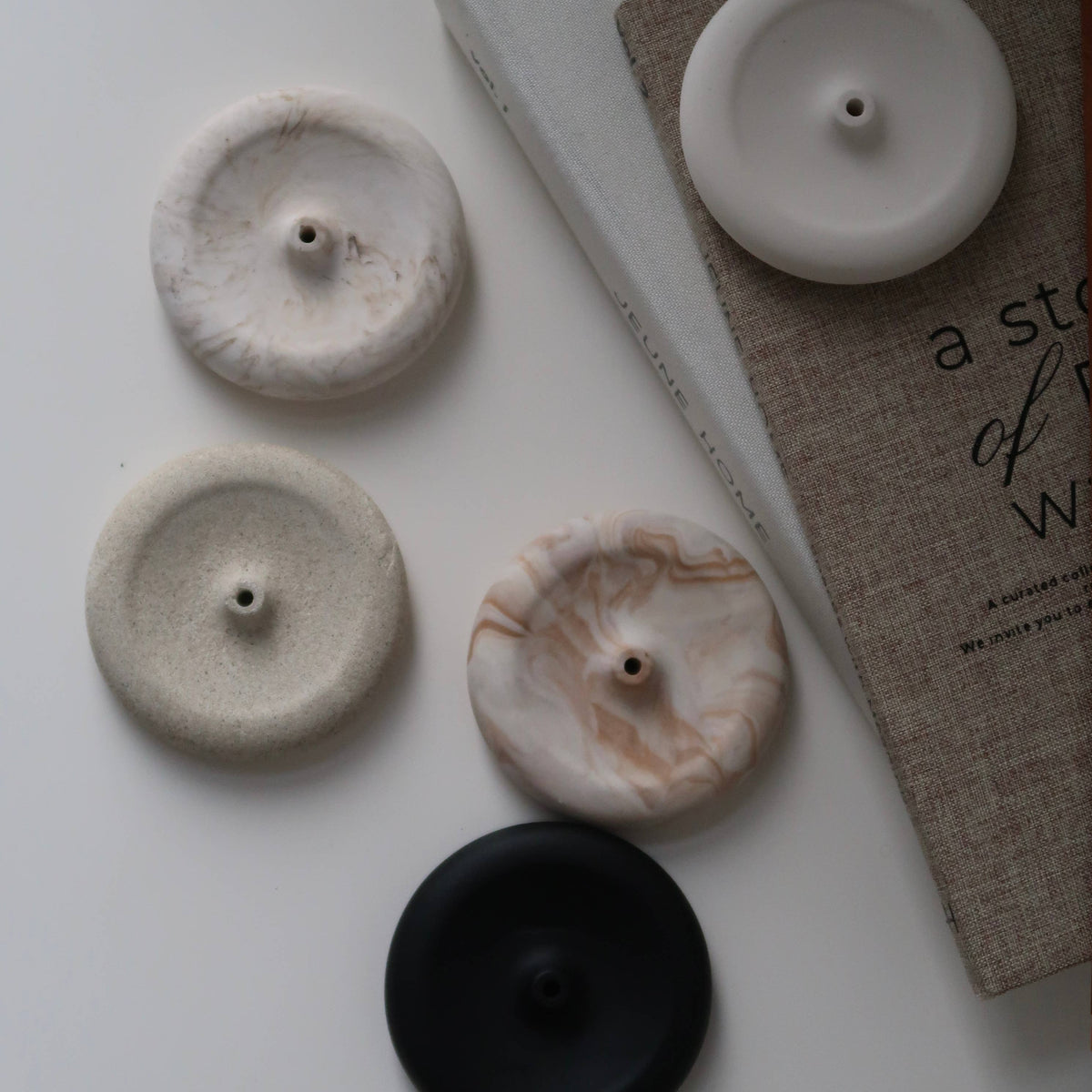 Four round button-like home decor pieces in white, cream, marbled beige, and black, arranged on a light surface beside a book.