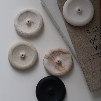 Four round button-like home decor pieces in white, cream, marbled beige, and black, arranged on a light surface beside a book.