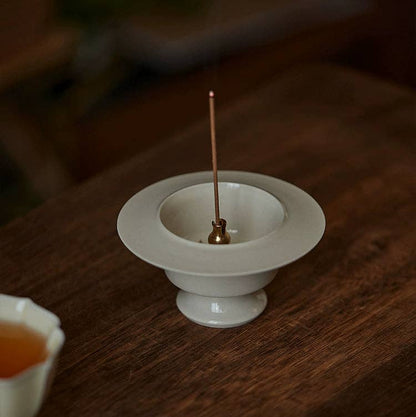 Incense burner with a single stick of incense on a wooden surface next to a small cup.