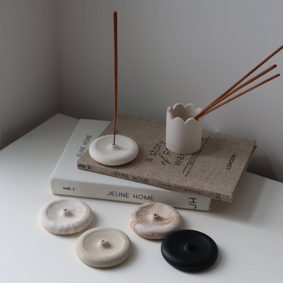 Minimalist ceramic incense holder and reed diffuser set arranged on stacked books with a few round incense burners in cream, beige, and black on a white surface against a light gray wall.