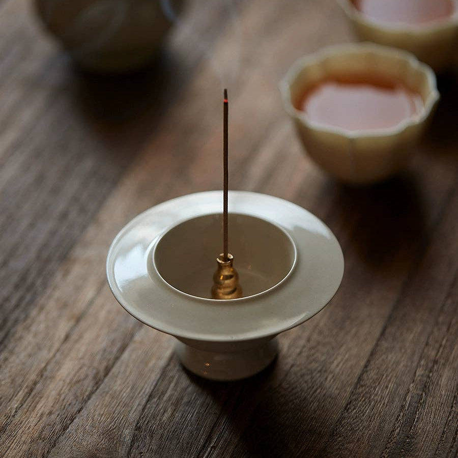 Incense stick on a small white dish with teacups and a teapot in the background on a wooden surface.