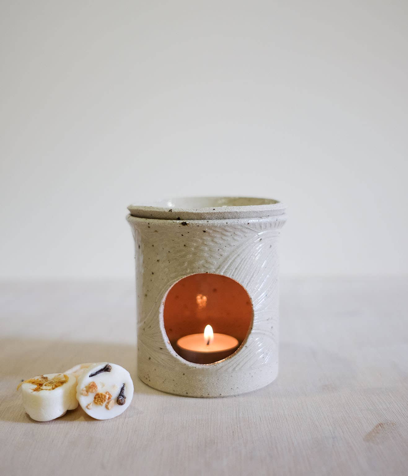 A speckled beige ceramic candle warmer with a lit tea light candle inside, shown on a light surface against a soft neutral background. Two small white wax melts are placed beside it.