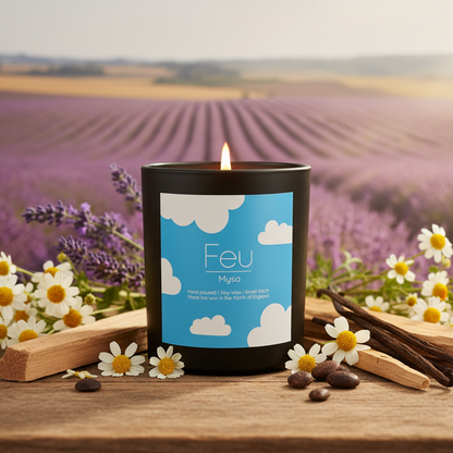 A black scented candle labeled 'Feu Myoï' sits on a wooden surface in a lavender field setting, surrounded by white daisies, lavender sprigs, cinnamon sticks, and coffee beans.