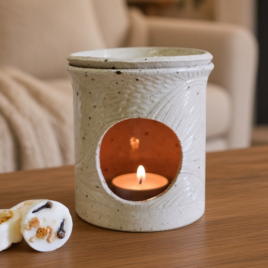 A ceramic candle warmer with a lit tea light candle sits on a wooden table in a cozy living room setting with a fireplace and sofa in the background. Two small wax melts are placed in the foreground.
