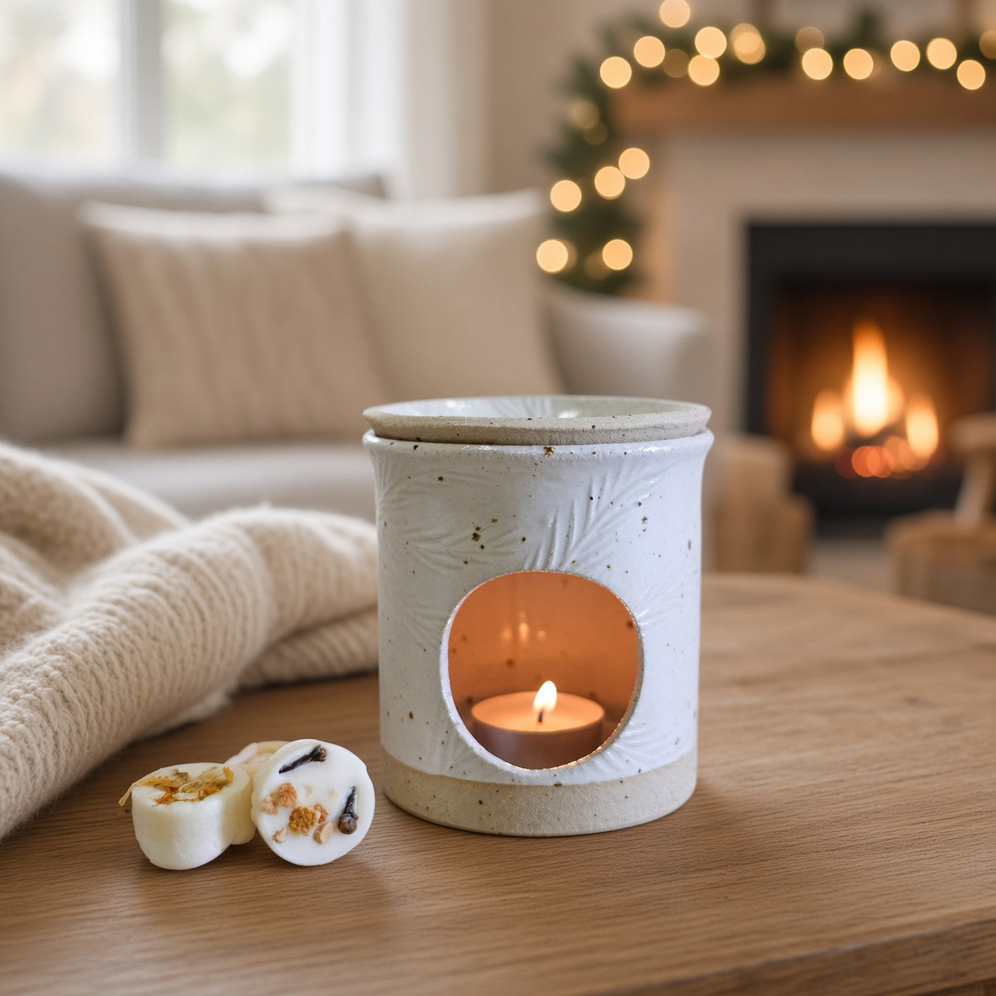 A ceramic candle warmer with a lit tea light candle sits on a wooden table in a cozy living room setting with a fireplace and warm bokeh lights in the background. Two small wax melts are placed beside it.