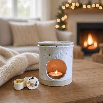 A ceramic candle warmer with a lit tea light candle sits on a wooden table in a cozy living room setting with a fireplace and warm bokeh lights in the background. Two small wax melts are placed beside it.