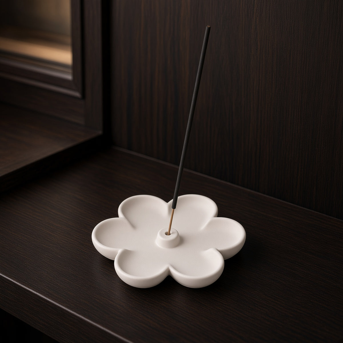 A white ceramic flower-shaped incense holder with a single incense stick inserted, set on a dark wooden surface against a dark wood-paneled background.