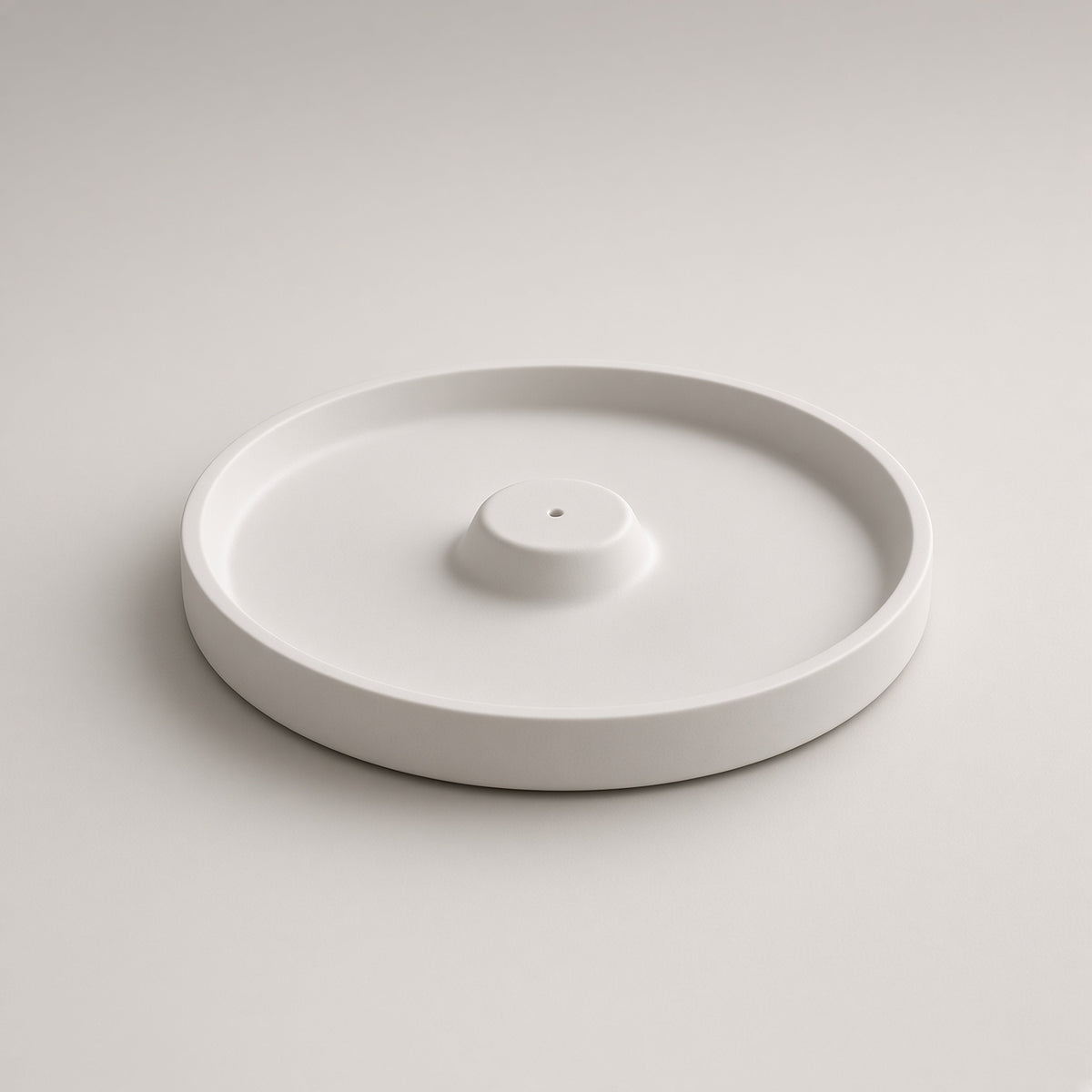 A minimalist white ceramic tray with a circular base and a small central pedestal, photographed on a clean light gray background.